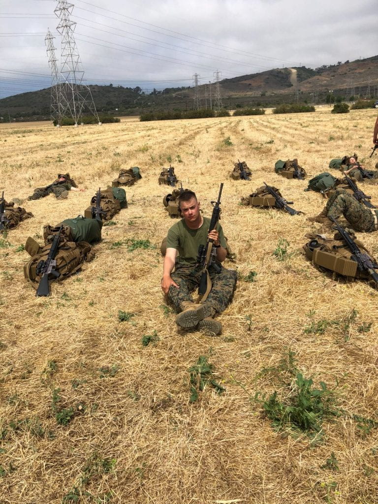 student sits in a field in military kit with packs laid around him
