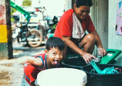 Little boy crouches over a bucket with his tongue out as presumably his mom works behind him washing something and smiling