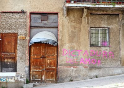 A bullet-riddled wooden door is on the left with a faded awning. In pink paint, there is graffiti on the wall to the right of the door reading, "Dictadura del amor"