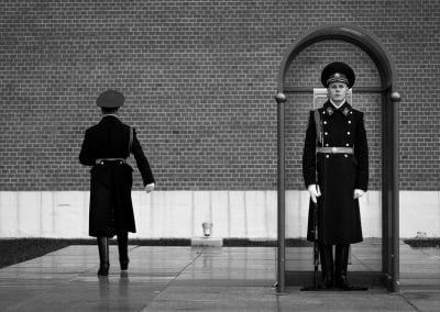 Black and white photo of two guards in front of a brick wall. One faces the camera as the other faces the wall.