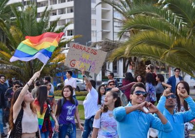 People in the streets surrounded by palm trees. One woman holding an LGBTQ flag, others forming heart shapes with their hands, another holding a sign reading, "Hugs from Mom: Free"