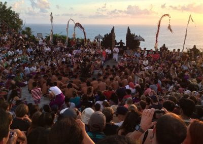 A large crowd of people in front of the ocean. Some are observing the dance and a group of shirtless men sit in the center performing.