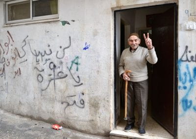 An elderly man holding a can and showing the peace sign with his left hand stands in a doorway of a plain white building covered in graffiti.