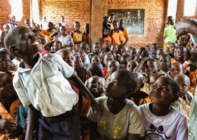 A group of children in a classroom are laughing and smiling at one boy who is standing, grinning towards the ceiling