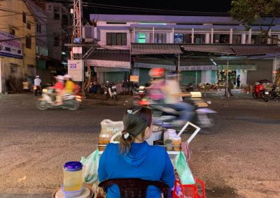 A woman vendor sits beside the road with her back to the camera. A motorcycle zooms past her in a blur.