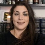 A headshot of Stacey Martino who is a white woman with long brown hair. The left side of her hair is behind her back while the right side is lying on top of her shoulder and is draped across her chest. She is wearing a black sweater and standing in front of a bookshelf smiling.