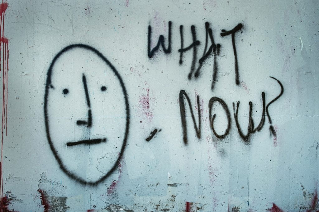 Graffiti on wall: Unsmiling face next to text "What now?"