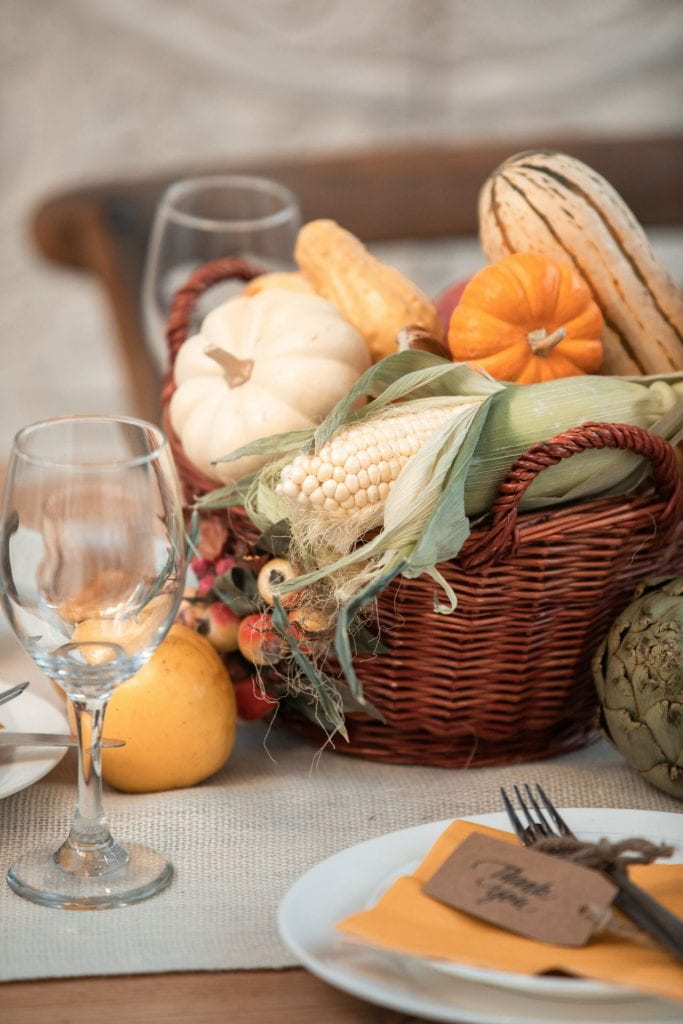 A photo of a table setting with two drinking glasses, a basket filled with corn and small gourds. There is a plate with a paper napkin and a fork on top. A brown paper tag with 'Thank You' written on it is tied to the fork. 