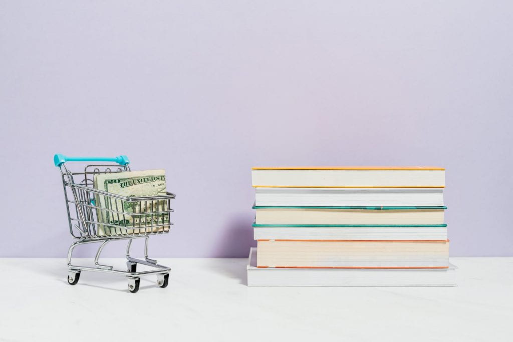 Image of shopping cart with $20 bills inside next to a stack of books.