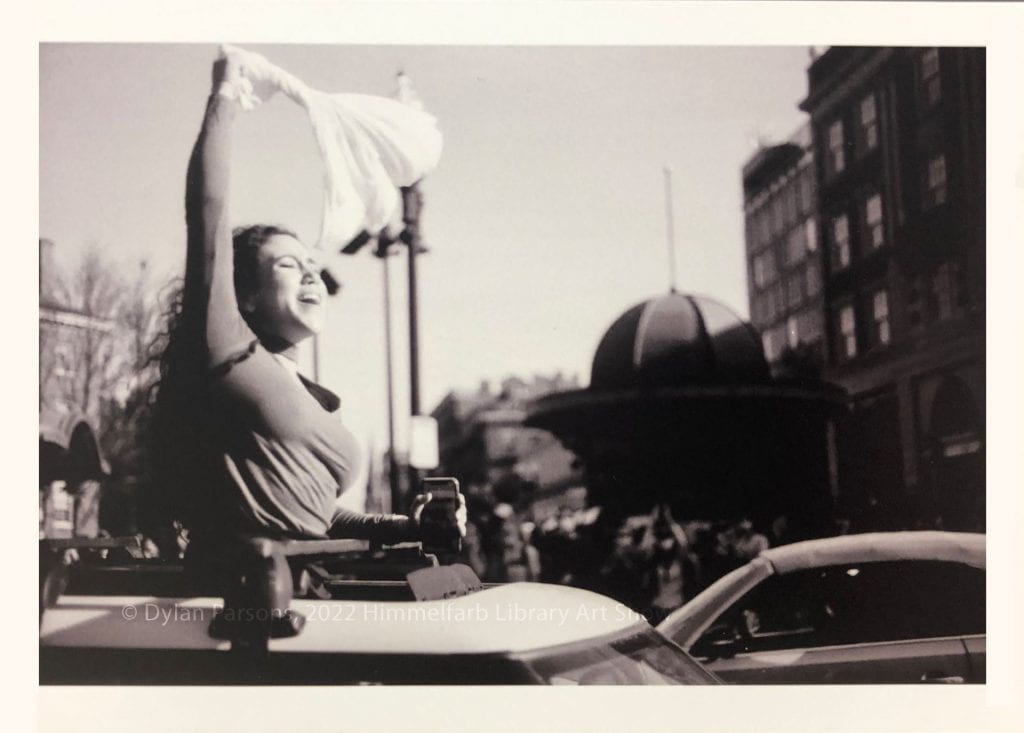 Black and white photograph of young woman standing up through a sun roof on a car waving a white shirt and smiling with eyes closed in celebration of Biden being elected President. 
