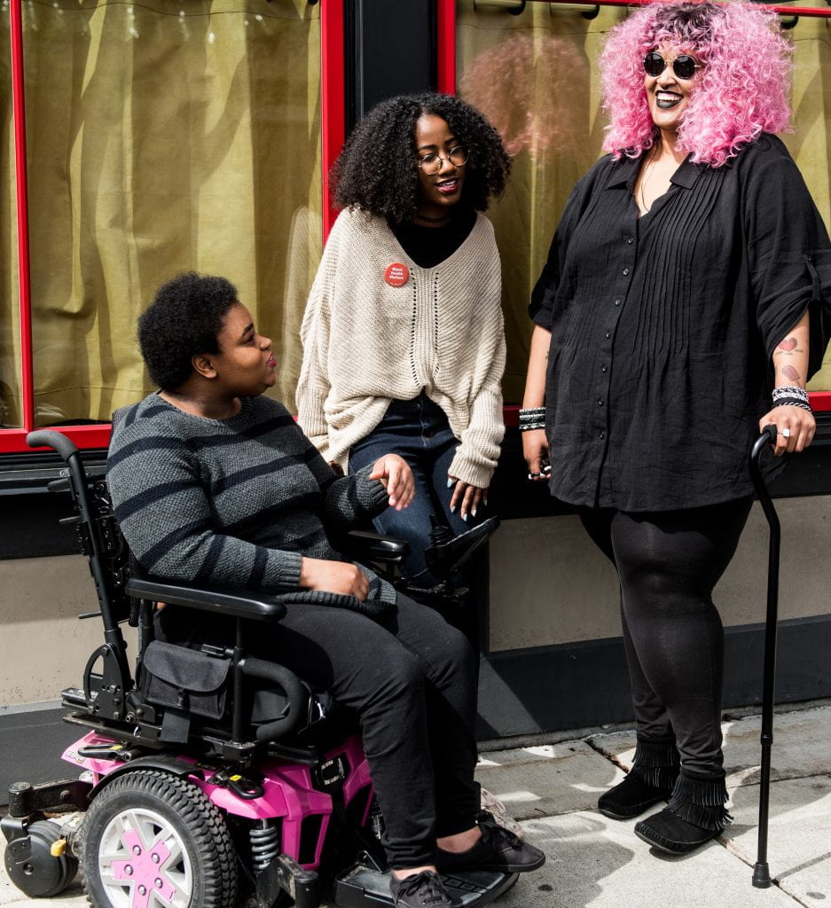 Photo of three individuals talking. One person is in a power chair, another leans against the wall and the third stands with their cane. 