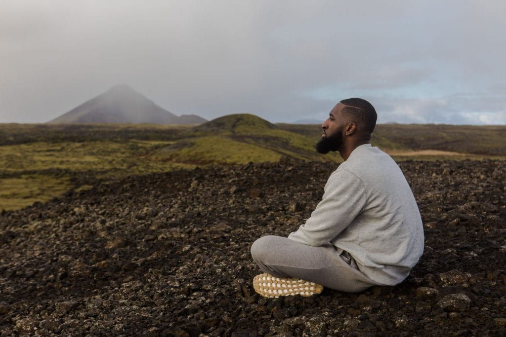 An African-American man dressed in a white shirt and grey pants is seated crossed legged on brown rocks and looks over a green, hilly landscape
