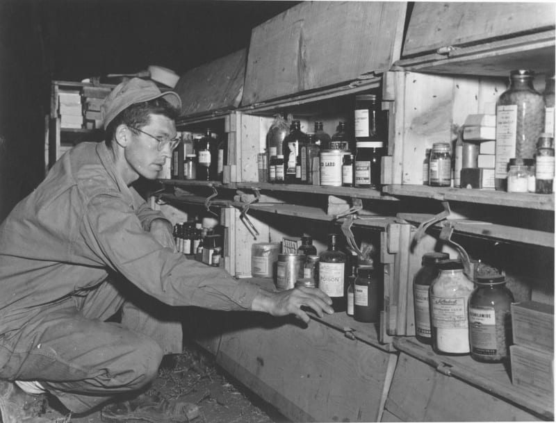 Black and white photo of a military medic kneeling in front of cabinet of medical supplies. 