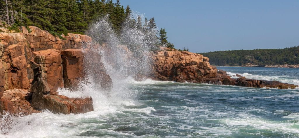 Picture of waves splashing against a coastline cliff with red rocks and pine trees above.