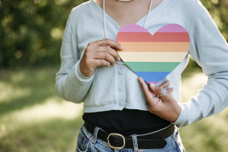 A person holding a cut out of a rainbow colored heart