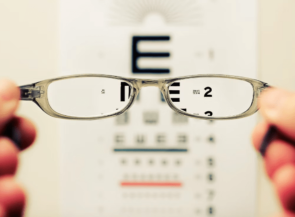 Picture of glasses being held in the foreground in front of a eye test chart that is blurred out in areas not covered by the glasses.
