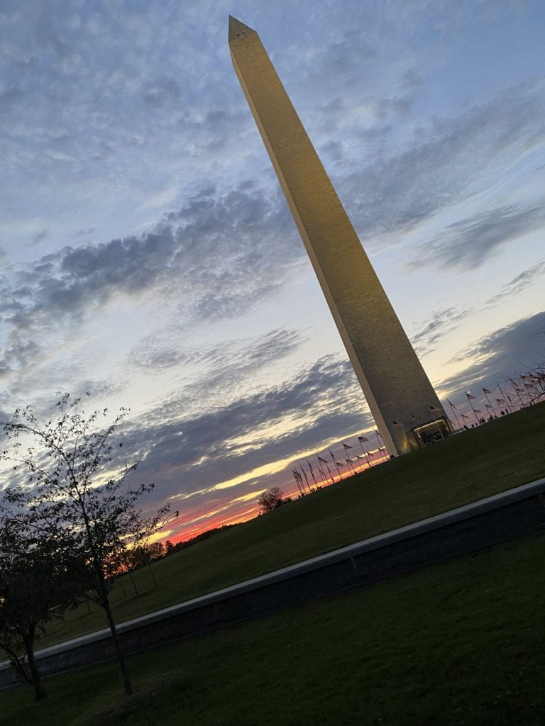 The Washington Monument with a scenic background of trees, grass, and a cloudy sky at sunset/sunrise. 