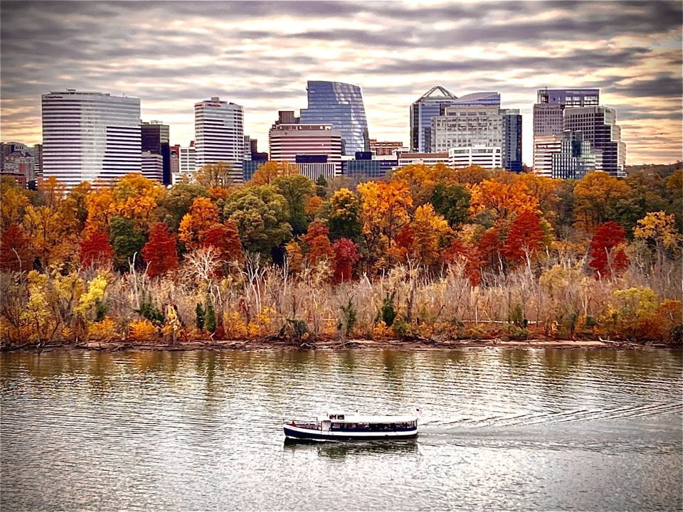 The image shows a boat floating on the water against a backdrop of a cloudy sky, colorful fall trees, and distant city skyline.