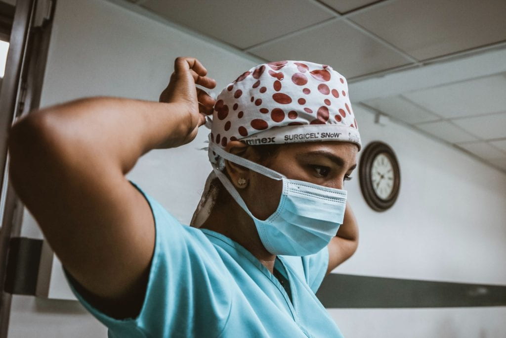 A person of color wearing blue scrubs and polka dotted surgical head covering secures a mask around their face.