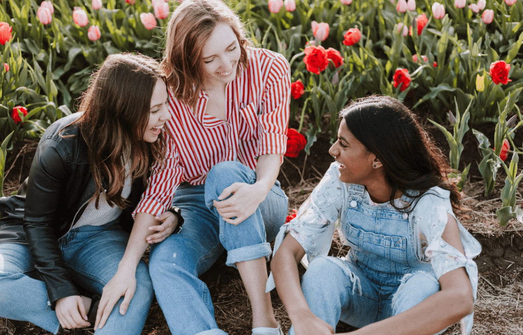 The image shows 3 women sitting in front of flowers. They are all wearing jeans and are sitting outdoors.