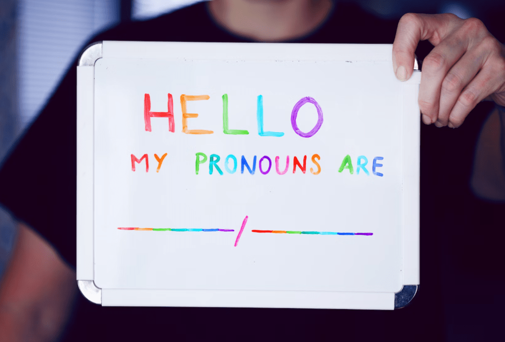 Picture of a person holding a white board with the text "Hello My Pronouns are ___" written in rainbow color letters.