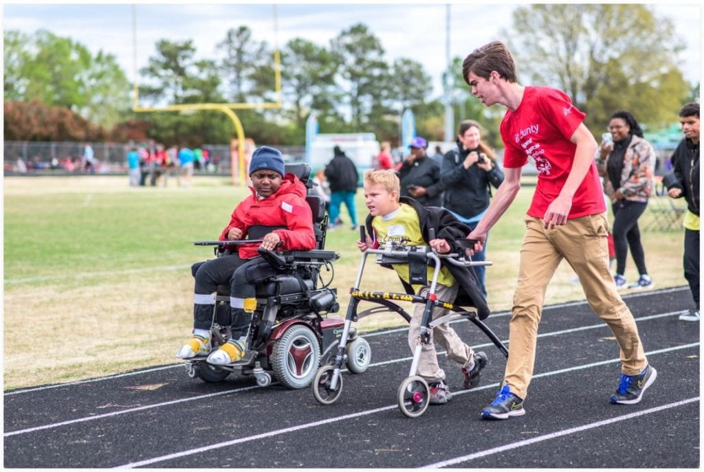 Child in a wheelchair and child using an assistive walking device are on a running track with a coach encouraging them