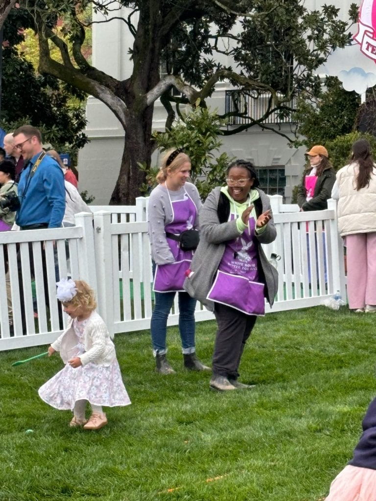 Two adults standing on a lawn looking on as children roll Easter eggs. One adult is applauding.