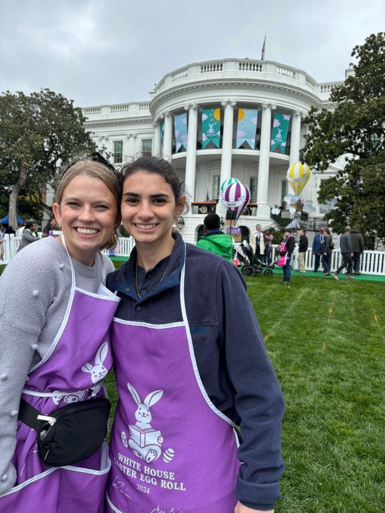 Two people stand in front of the White House with aprons that say White House Easter Egg Roll.