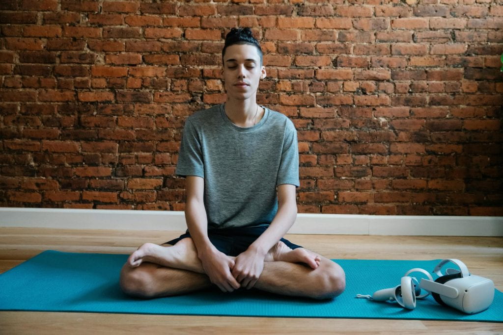 Picture of a person meditating in lotus pose on a yoga mat with a Virtual Reality headset nearby.