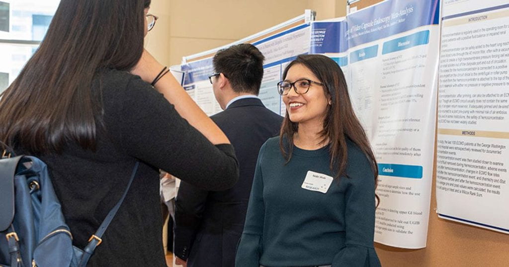 Picture of a student standing in front a research poster talking to another student.
